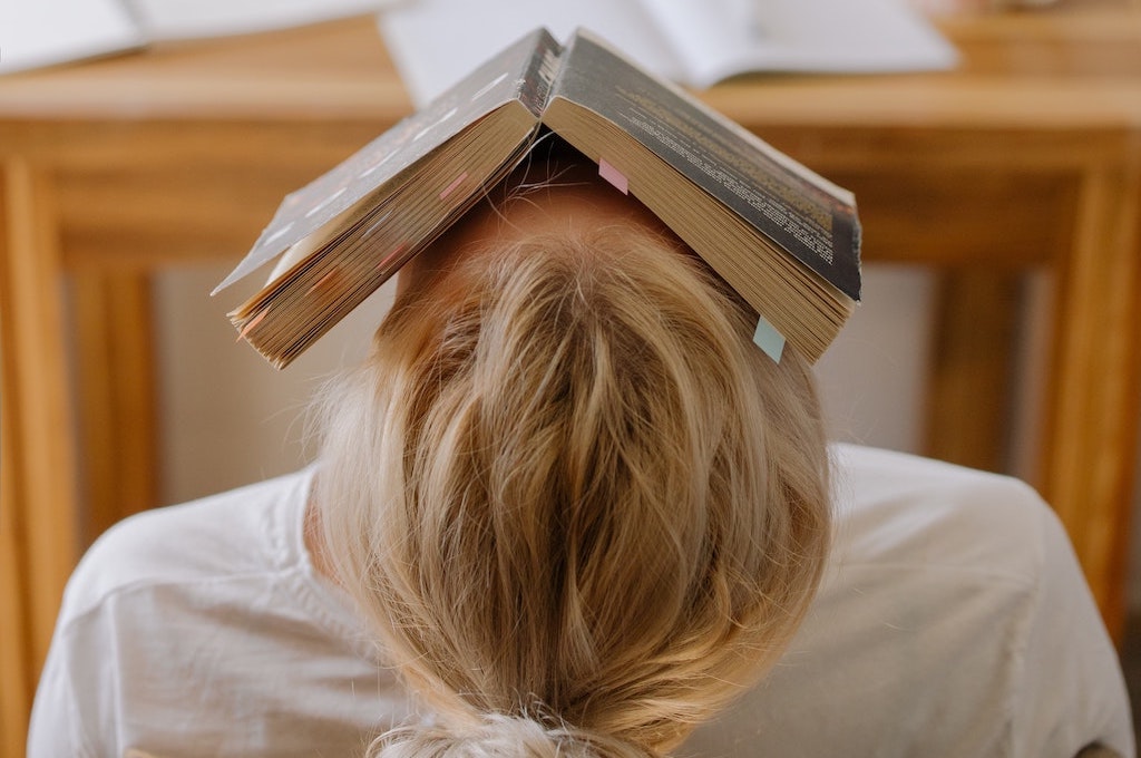 girl sitting in a chair at a wooden desk with a book opened on her face and leaning her head back