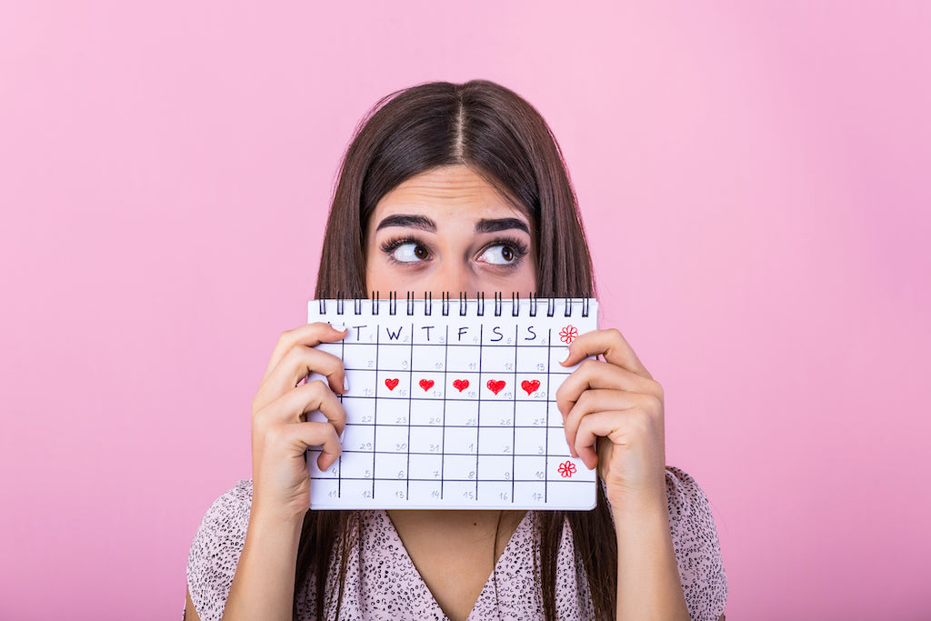 girl holding calendar in front of her face, with red hearts on 5 days of one week to imply her period cycle